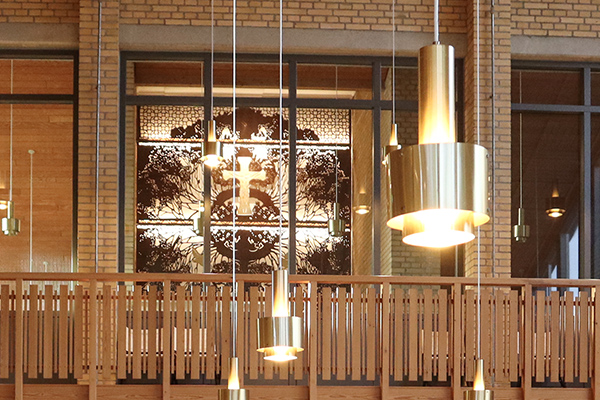 Hængende messinglamper foran murstensvæg og vinduer. Bagved skinner lys gennem et kunstværk i cortenstål.