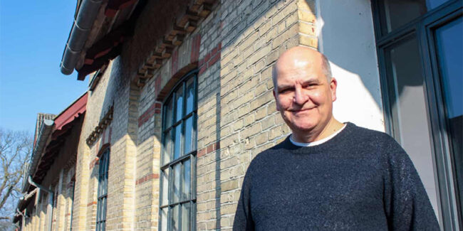 Thomas Hillerup i en mørkeblå sweater, der står foran en bygningsfacade (Materialgaarden) med mur af gule og røde mursten og med støbejernsvinduer.