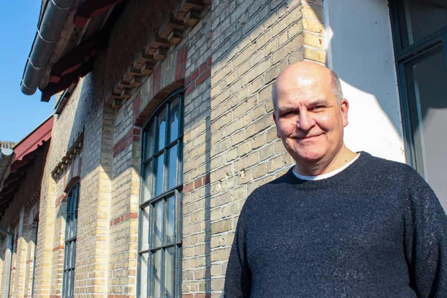Thomas Hillerup i en mørkeblå sweater, der står foran en bygningsfacade (Materialgaarden) med mur af gule og røde mursten og med støbejernsvinduer.