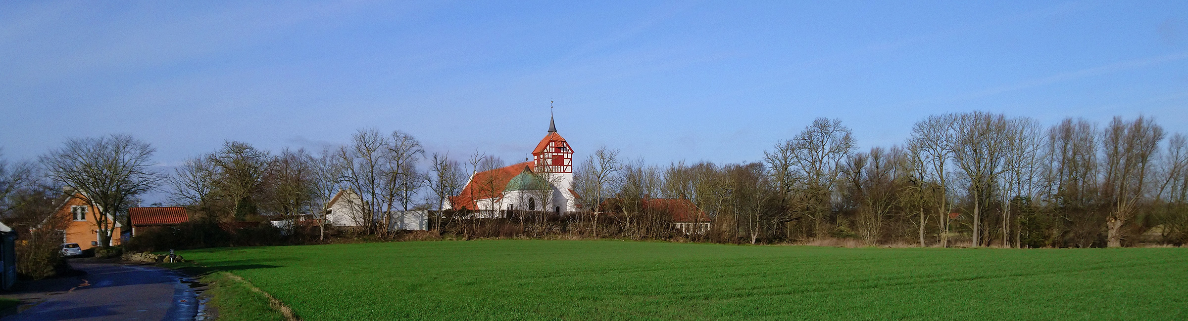 Viby Kirke set fra sydøst med blå himmel som baggrund og med grøn mark i forgrunden. Hvidkalkede vægge og nyt, rødt tegltag på skib og tårn.
