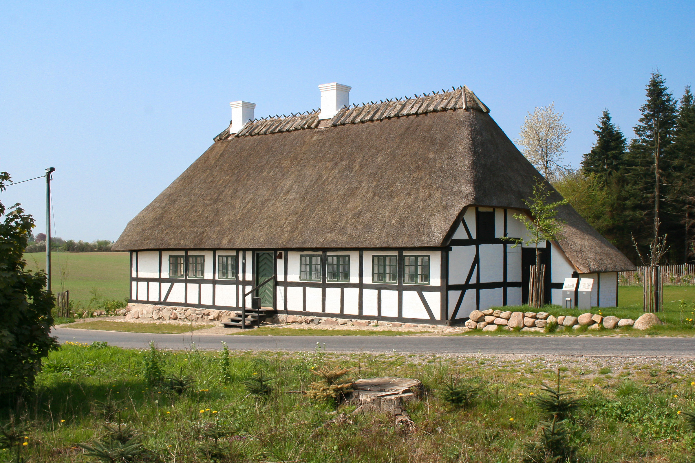 Bindingsværkshus med stråtag ved en vej med græs og træer omkring og blå himmel i baggrunden. Hvide tavl, sortopstreget tømmer og grønne døre og vinduer.