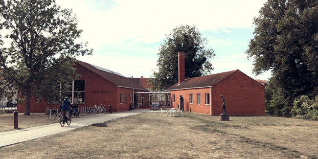 Ankomsten ved Nyborg Bibliotek. Rød murstensbygning med lavt saddeltag. Græsplæne og belagt sti foran. Træer omkring bygning. Hovedfløj til venstre og vestfløj til højre.