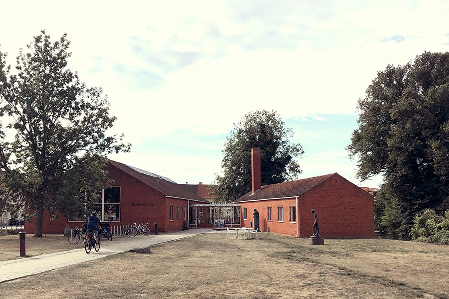 Ankomsten ved Nyborg Bibliotek. Rød murstensbygning med lavt saddeltag. Græsplæne og belagt sti foran. Træer omkring bygning. Hovedfløj til venstre og vestfløj til højre.