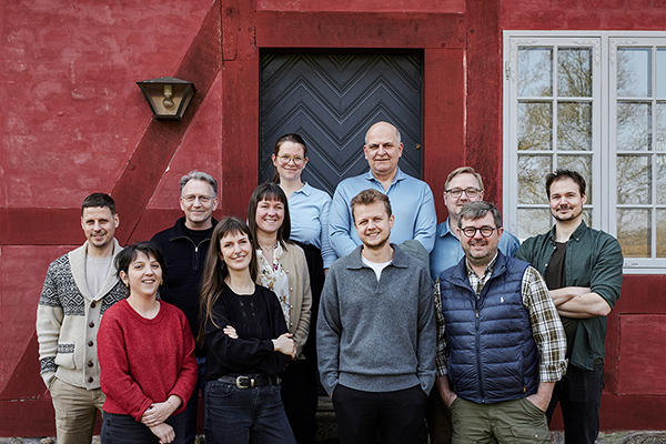 Gruppebillede af de ansatte hos Vaag Arkitekter. I alt 11 mænd og kvinder, der står foran en rødopstreget og rødkalket bindingsværksbygning med blå, flammeret dør og hvidt, småsprosset vindue.