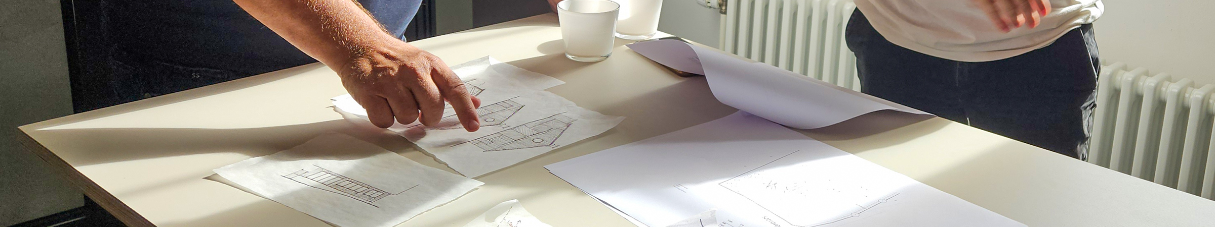 To personer står ved et bord og kigger på skitsetegninger. Den ene person peger på en tegning. Solen lyser ind fra siden på det lyse bord, hvor på der ligger flere ark.