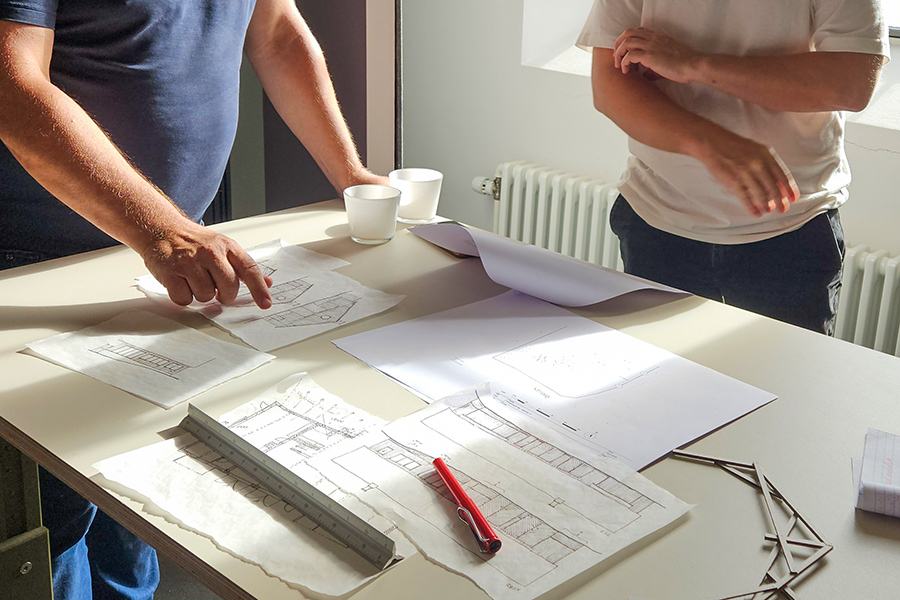 To personer står ved et bord og kigger på skitsetegninger. Den ene person peger på en tegning. Solen lyser ind fra siden på det lyse bord, hvor på der ligger flere ark, tegneredskaber m.v.